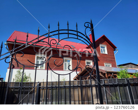 Wrought-iron gates on the background of a brown house with a red roof against the blue sky. Wrought-iron gates on the background of a brown house with a red roof against the blue sky. 123760126