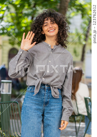 Young woman smiling friendly at camera, waving hands hello, hi, greeting or goodbye in city street 123762612