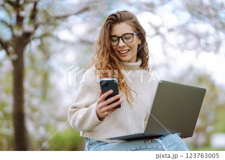 Happy woman with laptop and phone in spring blooming park while working online. Freelancing concept. 123763005