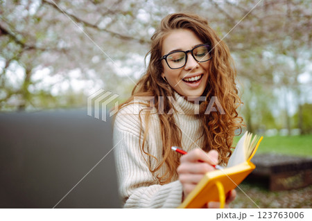 Woman sits on green grass near flowering trees, with laptop, writes in notepad. Planning concept 123763006