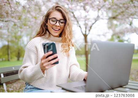 Happy woman with laptop and phone in spring blooming park while working online. Freelancing concept. Happy woman with laptop and phone in spring blooming park while working online. Freelancing concept. 123763008