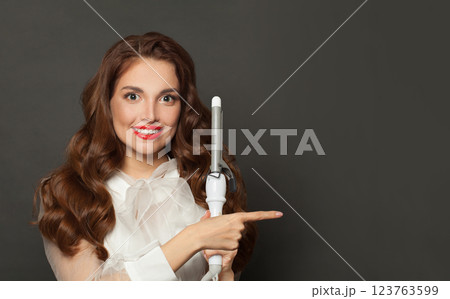 Elegant brown haired woman with shiny long curly hairstyle showing curling iron. 123763599
