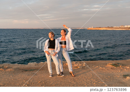 Women Beach Sunset Pose - Two women stand on a rocky cliff overlooking the ocean at sunset. They are both smiling and looking at the camera, one has her arm raised in the air. Women Beach Sunset Pose - Two women stand on a rocky cliff overlooking the ocean at sunset. They are both smiling and looking at the camera, one has her arm raised in the air. 123763896