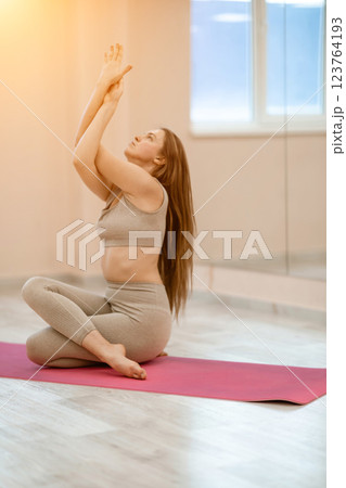 Young woman doing yoga in the gym. A girl with long hair and in a beige tracksuit stands in a cow pose on a pink carpet. A woman performs Gomukhasana. 123764193