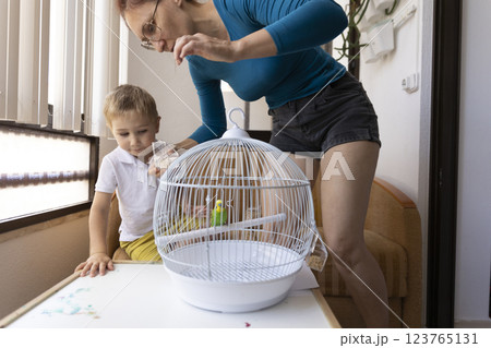 Mother and son feeding a small parrot in a cage at home 123765131