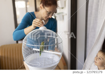 Woman closing birdcage with green parrot inside, child observing Woman closing birdcage with green parrot inside, child observing 123765179