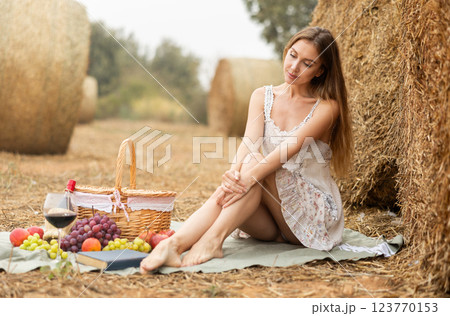 Young pretty woman in white dress sitting on soft mat near stack of straw with basket of fruit and wine, picnic in field Young pretty woman in white dress sitting on soft mat near stack of straw with basket of fruit and wine, picnic in field 123770153