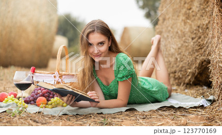 Young smiling sexy woman wearing in summer green dress lying down and reading book on soft mat near with stack of straw and enjoying romantic picnic 123770169