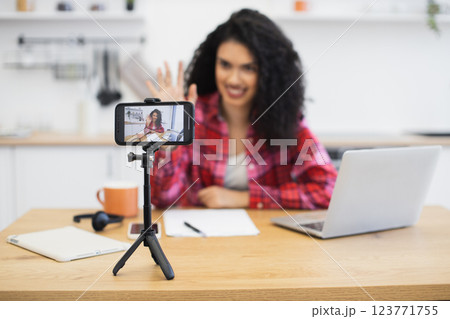 Young African woman filming content with smartphone, laptop, and stationery in kitchen setting. Creates social media content using technology at home. Young African woman filming content with smartphone, laptop, and stationery in kitchen setting. Creates social media content using technology at home. 123771755