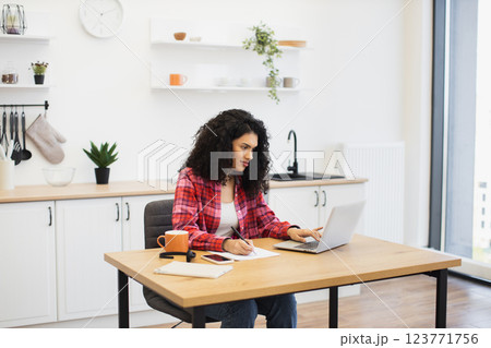 African American young adult female wearing red checked shirt working on laptop at home. Comfortable kitchen environment, focused on remote work productivity. 123771756