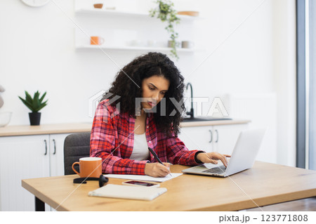 Young African American woman wearing a red plaid shirt writing and working on laptop, seated in bright, modern home office, focusing on education or remote work. 123771808
