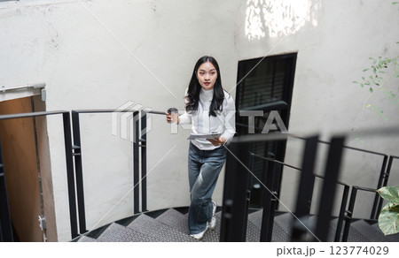 Young Asian woman walking up spiral staircase with coffee and documents, balancing work and daily routine. 123774029