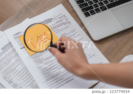 Hand examining tax documents with a magnifying glass on a wooden desk. Hand examining tax documents with a magnifying glass on a wooden desk. 123774034