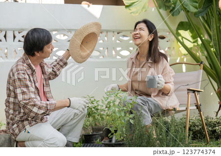 Diverse couple laughing together while gardening, sharing a hat and a cup. 123774374