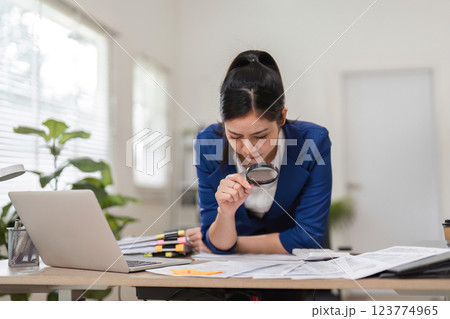 Accountant scrutinizing financial papers with a magnifying glass in a contemporary workspace. 123774965