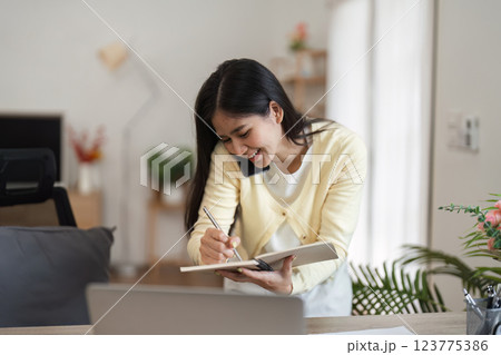 Smiling woman engaged in a phone call while writing in a notebook at her home office. 123775386