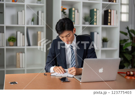 Young professional in suit taking notes while working on laptop, focused on tasks in modern office environment. 123775403