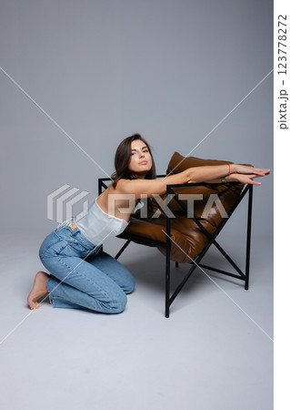 Fashion model kneeling and posing with a modern chair in studio 123778272