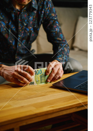 Man holding one hundred euro banknotes, sitting on sofa in front of table with open laptop. Person counting cash money at home. Personal finance and home budget concept Man holding one hundred euro banknotes, sitting on sofa in front of table with open laptop. Person counting cash money at home. Personal finance and home budget concept 123779343