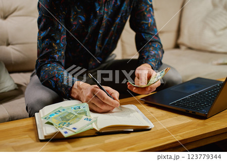 Man managing finances, writing in notebook while holding euro banknotes. Concept of budgeting, saving and financial planning 123779344