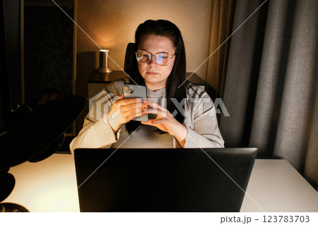 Serious woman freelancer with eyeglasses using smartphone while working with laptop. 123783703