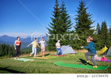 Group of people doing yoga pose outdoor in camping in the mountains. Adults and children standing on yoga mats, each doing a yoga pose under a clear blue sky in the morning. Young boy is instructor. 123784476