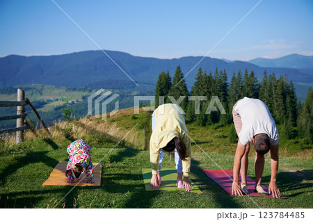 Young family doing yoga outdoors in scenic mountainous landscape in morning. Father, mother and daughter on yoga mats, with scenic view of mountains and forest in background under clear blue sky. Young family doing yoga outdoors in scenic mountainous landscape in morning. Father, mother and daughter on yoga mats, with scenic view of mountains and forest in background under clear blue sky. 123784485