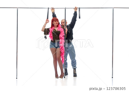 Woman in pink boa standing confidently as man in black leans in behind gripping rail against white studio background. Concept of street style 123786305
