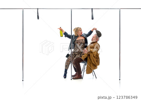 Woman teasing elderly man while holding bottle, pulling his cap against white studio background. 123786349