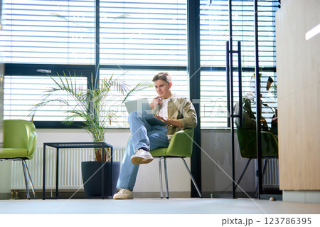 Thoughtful young man, dressed casually, analyzing data on laptop in bright office with contemporary decor and sleek furniture. 123786395