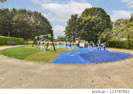 A vibrant playground features swings and play equipment surrounded by lush trees and a blue safety surface, perfect for children's outdoor fun. 123787662