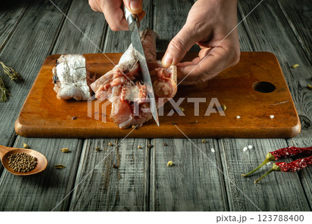 In a cozy kitchen, a skilled chef carefully cuts raw hake on a weathered wooden cutting board. Spices and herbs are scattered throughout, evoking a sense of culinary creativity and tradition 123788400