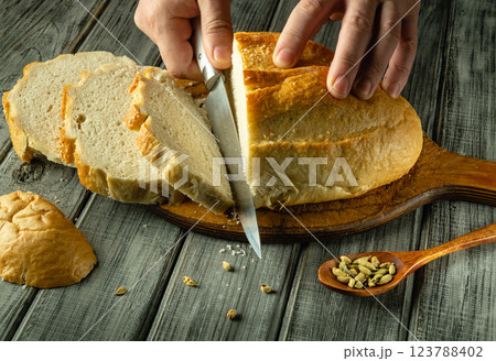 Warm bread rests on a wooden board, as hands meticulously slice through its crusty exterior. Sunlight bathes the setting, revealing seeds scattered nearby 123788402