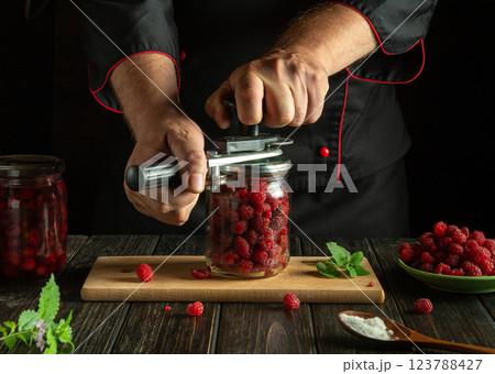Preparing canned raspberries with mint. A cook rolls up a jar of raspberries with a hand seaming key on a kitchen board. Low key concept of making raspberry compote 123788427