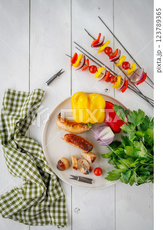 Grilled sausages and vegetable skewers with vegetables and herbs on a light background. View from above Grilled sausages and vegetable skewers with vegetables and herbs on a light background. View from above 123789365