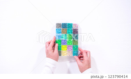 Flat lay. Woman hands gracefully poised over a collection of beads, sorted by color in transparent organizers. The array of beads spans a vibrant spectrum, from deep purples to bright oranges 123790414