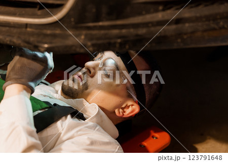 Focused Auto Mechanic Inspecting Vehicle Underneath Using Safety Equipment in Garage Workshop Focused Auto Mechanic Inspecting Vehicle Underneath Using Safety Equipment in Garage Workshop 123791648