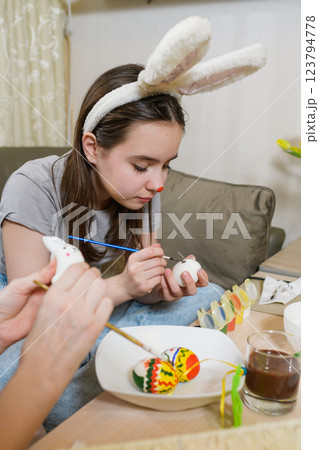 A young girl wearing bunny ears paints a white Easter egg with a brush. 123794778