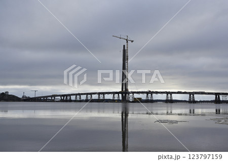 A large-scale bridge construction project stretches across a calm body of water, with a towering central pylon and a crane reaching skyward A large-scale bridge construction project stretches across a calm body of water, with a towering central pylon and a crane reaching skyward 123797159