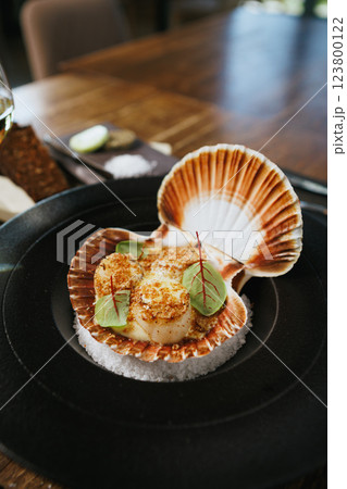 Grilled scallops in a shell with a crispy breadcrumb topping, adorned with fresh microgreens. Gourmet seafood appetizer presented on a black plate with a bed of salt Grilled scallops in a shell with a crispy breadcrumb topping, adorned with fresh microgreens. Gourmet seafood appetizer presented on a black plate with a bed of salt 123800122