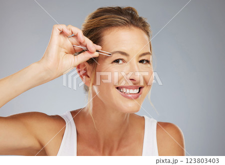 Face, eyebrows and senior woman plucking hair for beauty, wellness and clean body against a grey mockup studio background. Portrait of an elderly model doing skincare with tweezers and mock up space 123803403