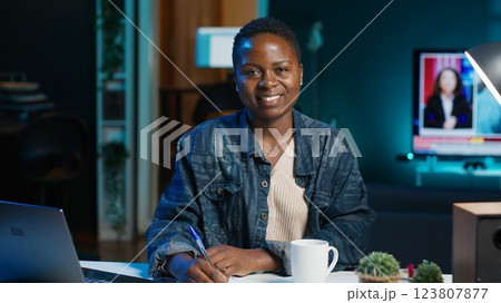 Portrait of smiling woman sitting on chair at home office desk, writing notes on notebook. Cheerful person in living room transcribing information from laptop screen on paper, camera A 123807877