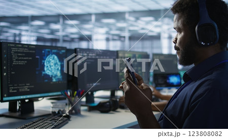 Worker in data center wearing headphones taking break from implementing AI systems, using smartphone. Focus on artificial intelligence server farm employee using mobile phone, camera A 123808082