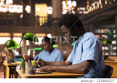 Black med student in scrubs does research and takes notes for his thesis, working hard at the campus library to become a doctor and obtain his diploma. Young man studying from copybooks. 123808217