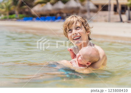 An older brother gently holds his baby sibling while swimming in the sea, sharing a joyful and heartwarming moment of family bonding and summer fun in the water. A perfect blend of love, care, and 123808410