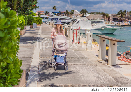 A young mother walks along the scenic waterfront, pushing a baby stroller with her child inside. A peaceful and heartwarming moment of motherhood, relaxation, and outdoor bonding by the sea 123808411