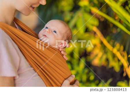 A loving mother walks through a peaceful park, carrying her baby securely in a sling. A heartwarming moment of maternal bonding, babywearing, and the joy of motherhood in nature 123808413