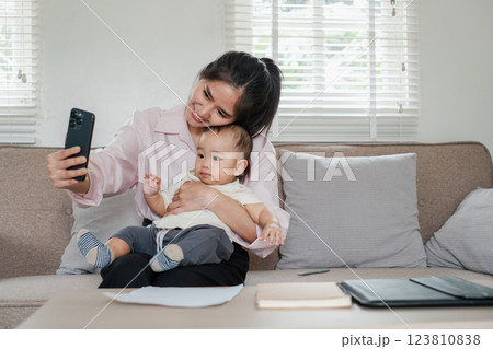 A joyful mother and her baby capturing a selfie together on a comfortable sofa in a well-lit living room. A joyful mother and her baby capturing a selfie together on a comfortable sofa in a well-lit living room. 123810838