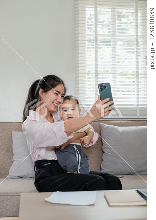 A joyful mother and her baby capture a selfie together on a comfortable sofa in a well-lit living room. 123810839