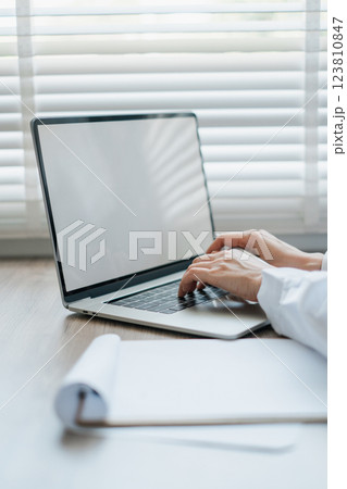 Close-up of hands typing on a laptop in a sunlit office setting, with blinds and a notepad nearby. 123810847